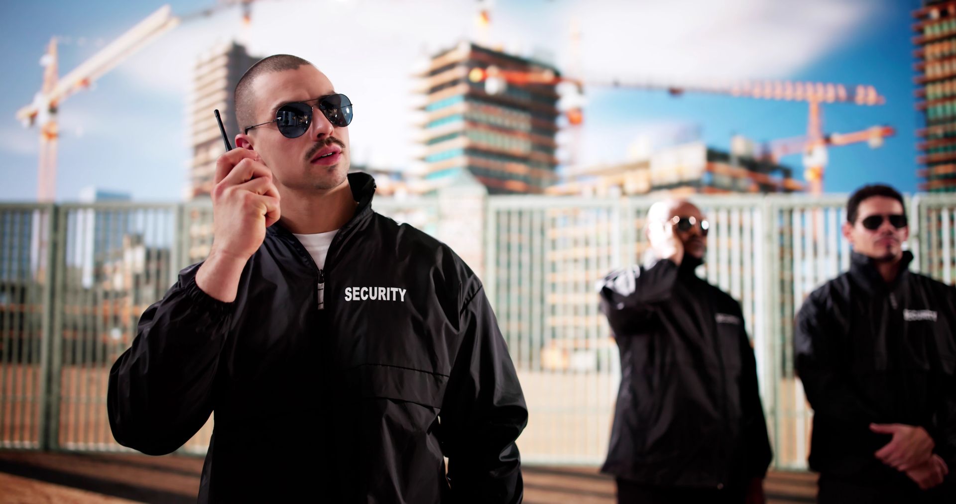 Security guards in black uniforms and sunglasses stand outdoors near construction cranes.