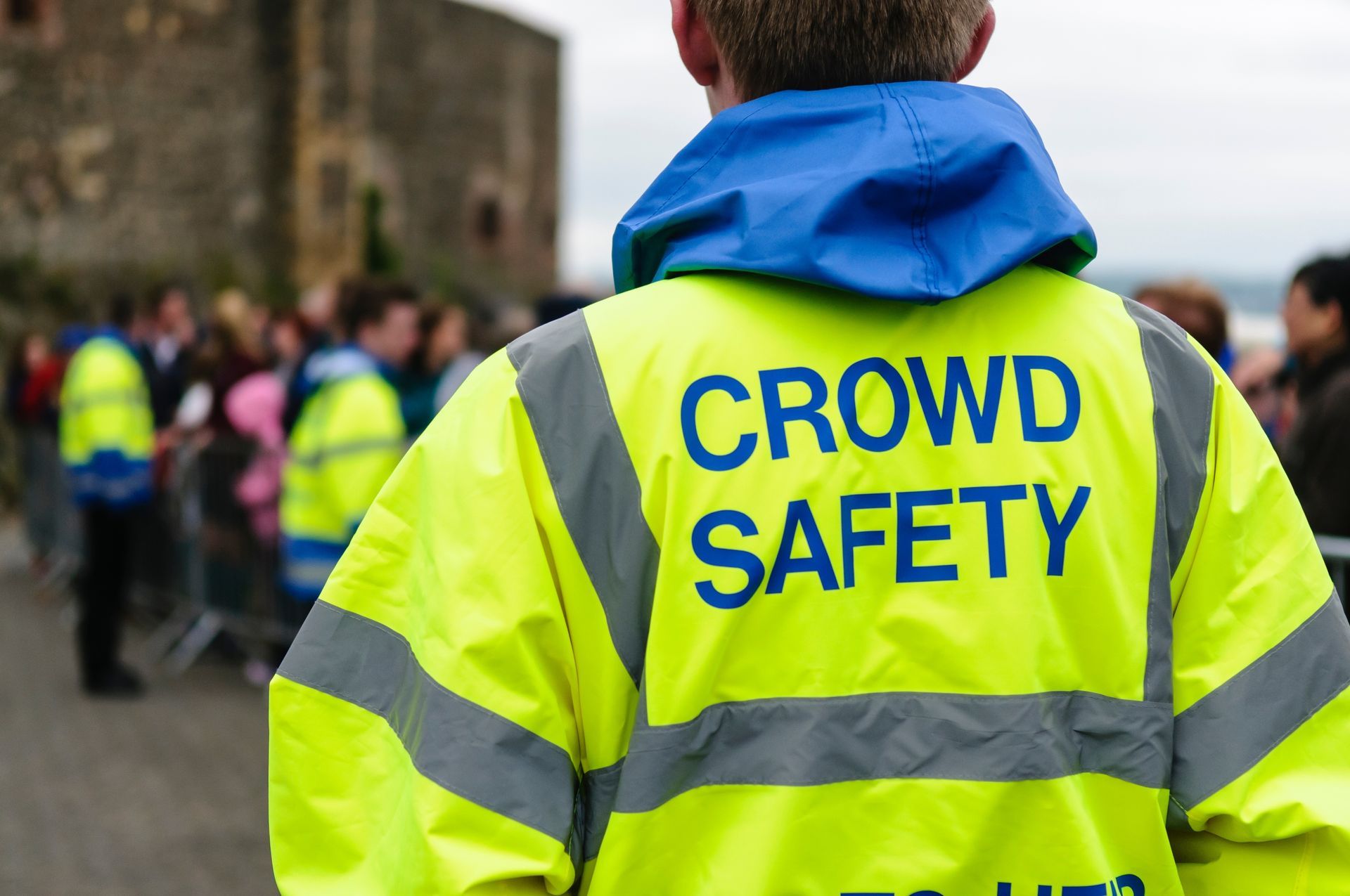 Crowd safety worker in a fluorescent yellow jacket with blue hood, standing outdoors at an event