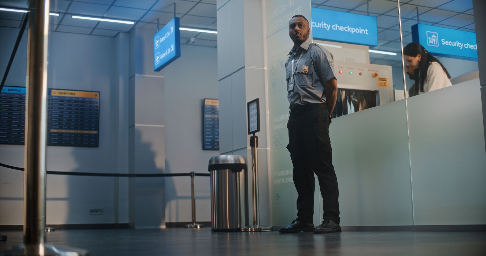 Airport security checkpoint with an officer standing by a counter and a traveler behind the glass partition.