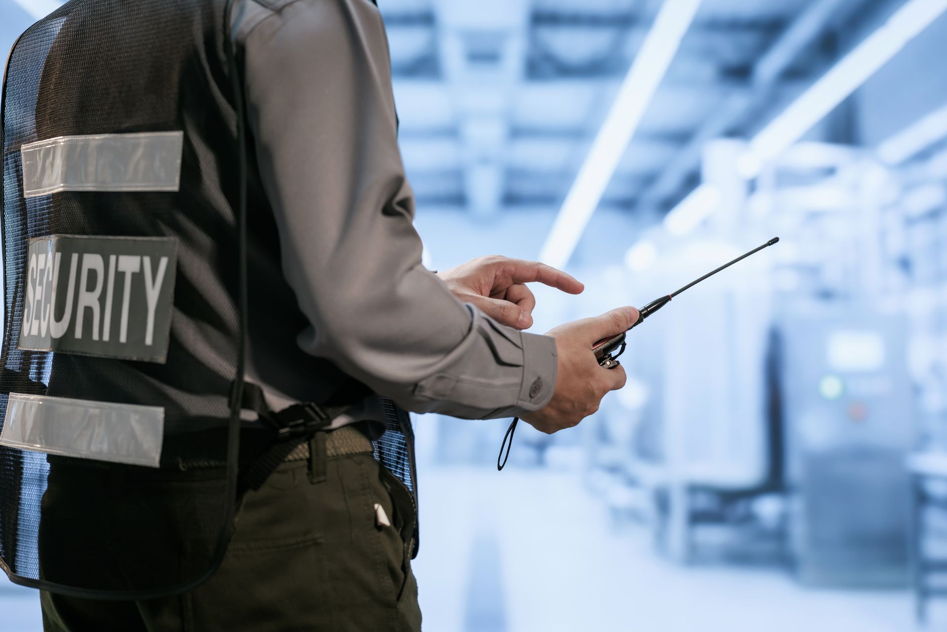 Security guard holding a walkie-talkie in an industrial hallway, wearing a reflective vest labeled “SECURITY.”