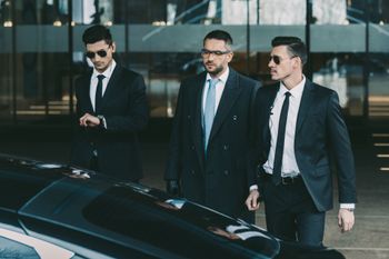 Three suited professionals walking beside a black car outside a glass building.