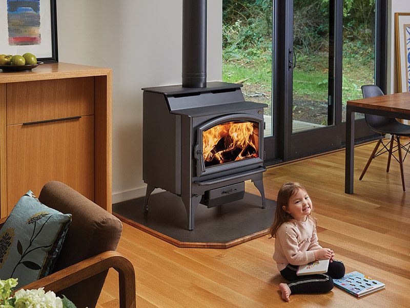 A little girl is sitting on the floor in front of a wood stove.