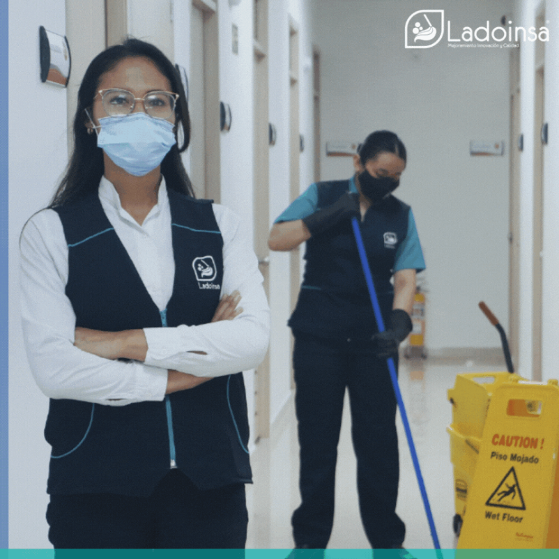 Una mujer con una máscara está parada en un pasillo junto a un balde amarillo de precaución de piso mojado.