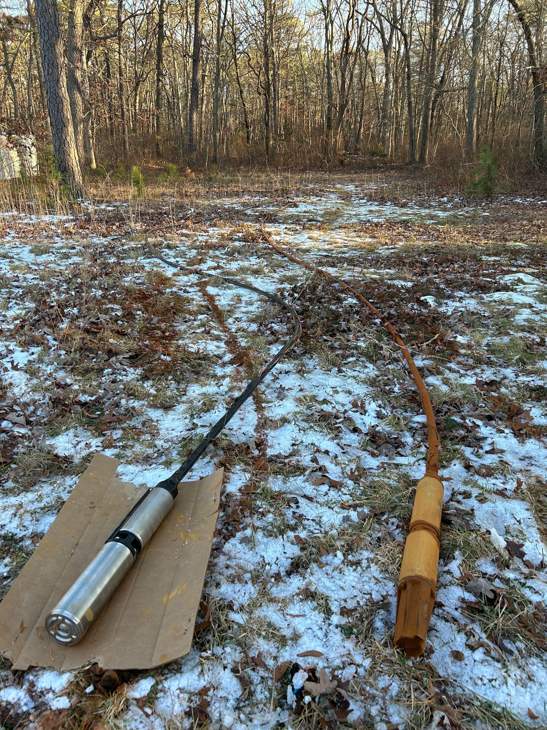 Two dowsing rods on snowy ground in a wooded area. One is metal, the other wood. Brown leaves and trees are visible.