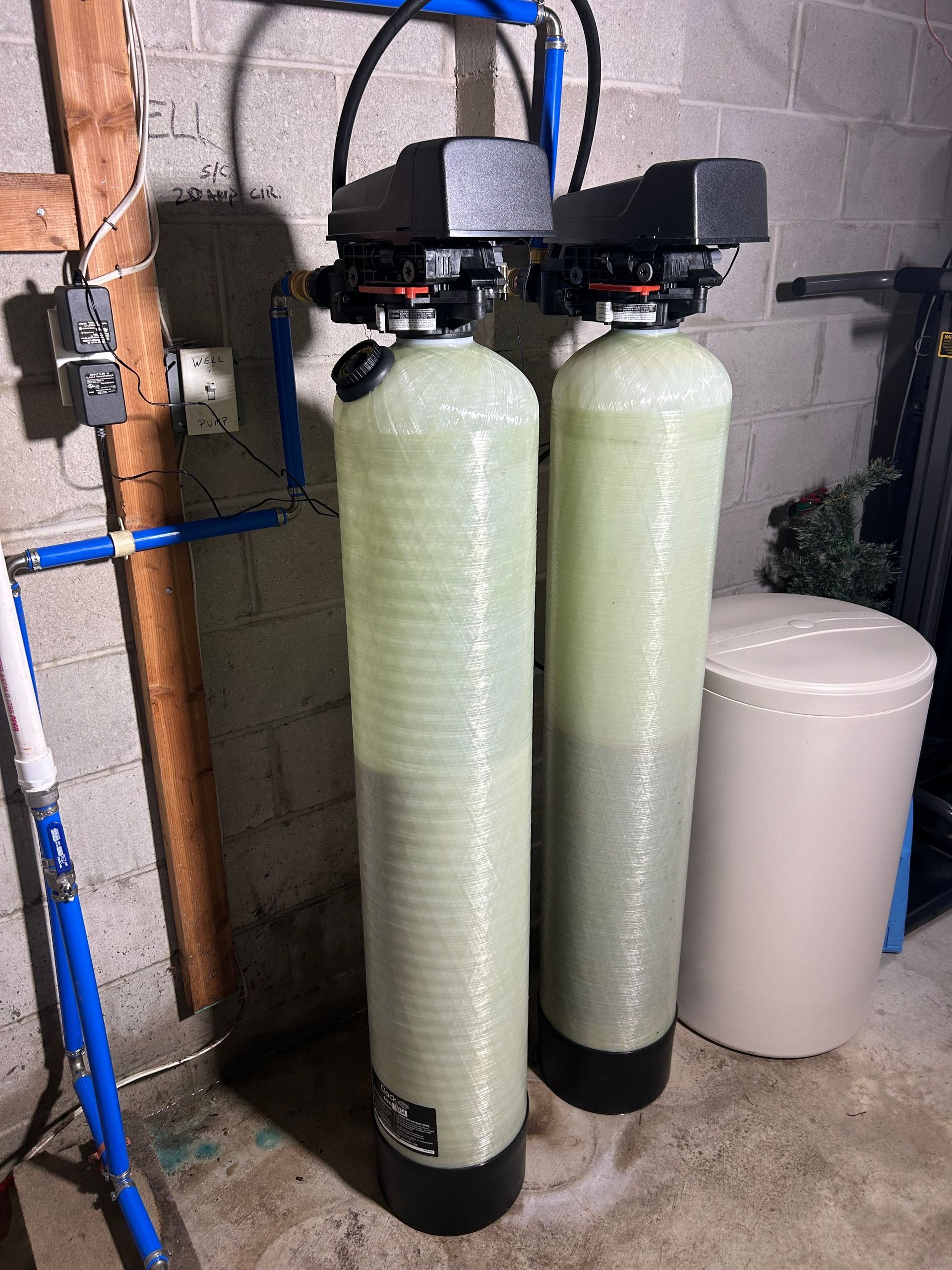 Two tall water filtration tanks with black control heads, pipes, and a white bin in a basement.