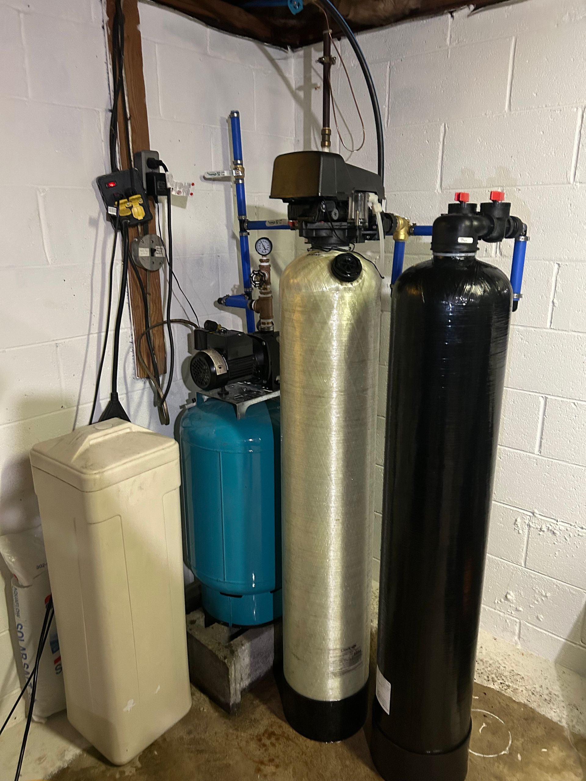 Water filtration and softening system in a basement. Tall tanks are silver and black, with a blue pressure tank.
