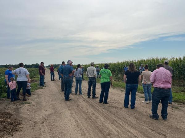 group of people standing on a dirt road listening to a speaker next to a corn field