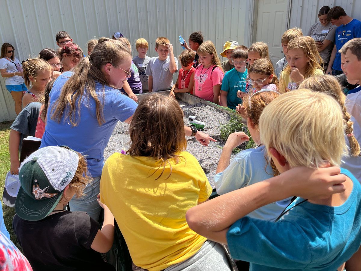group of children listening to a woman showing them items on an outdoor table