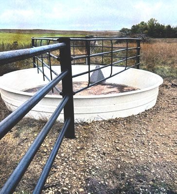 A large, round, white livestock water tank sitting on dry, gravelly ground in an open field.
