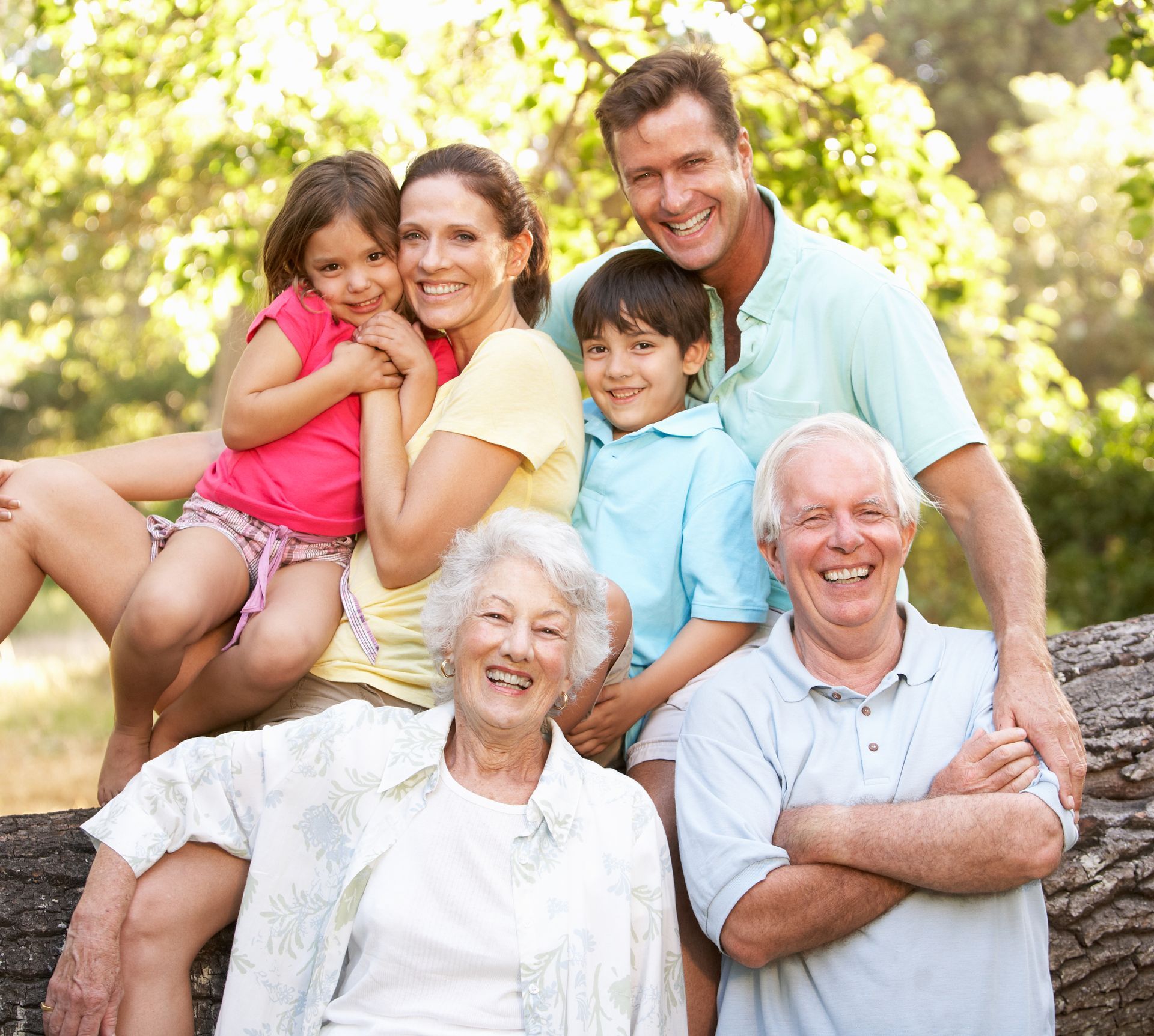 Family of seven smiling outdoors, including grandparents, parents, and children; sunny setting.