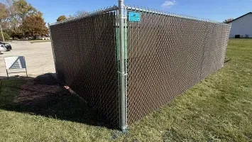 Chain-link fence corner with brown privacy mesh beside a grassy field and sidewalk.