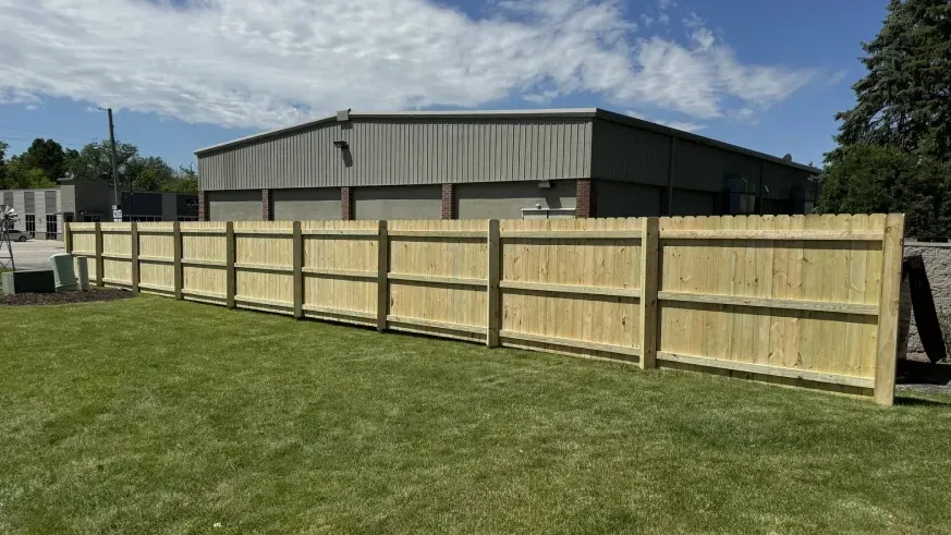 Wooden privacy fence along a grassy yard beside a gray metal building under a partly cloudy sky.