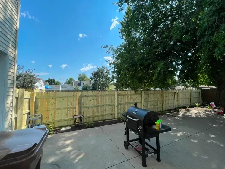 Backyard patio with a black grill, wooden fence, and trees under a bright blue sky