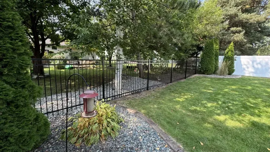 Black metal fence beside a grassy yard and trees, with a small red birdhouse or lantern in the foreground