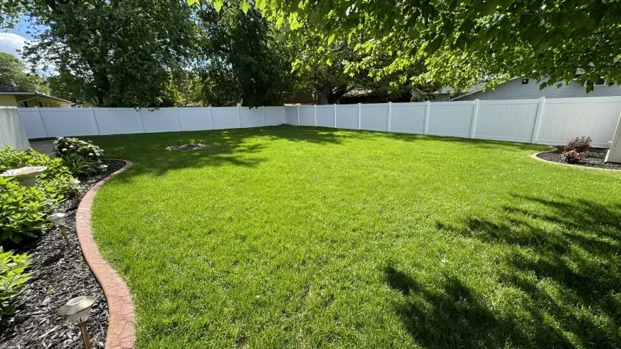 Sunny backyard with a lush green lawn, white fence, and garden beds along the edges