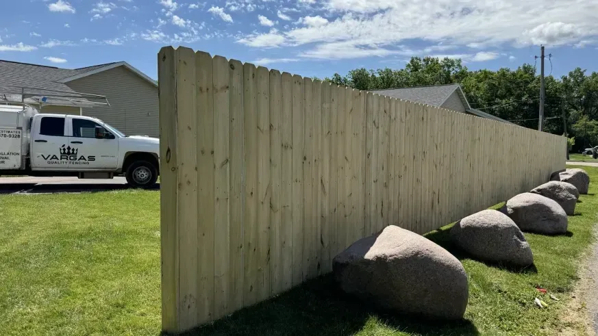 Long wooden fence in a grassy yard with large rocks, beside a white work truck and house under a blue sky