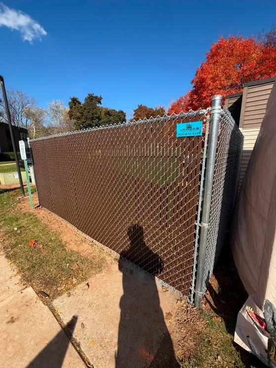 Chain-link fence along a sidewalk with a turquoise sign, autumn tree, and blue sky; person’s shadow in foreground