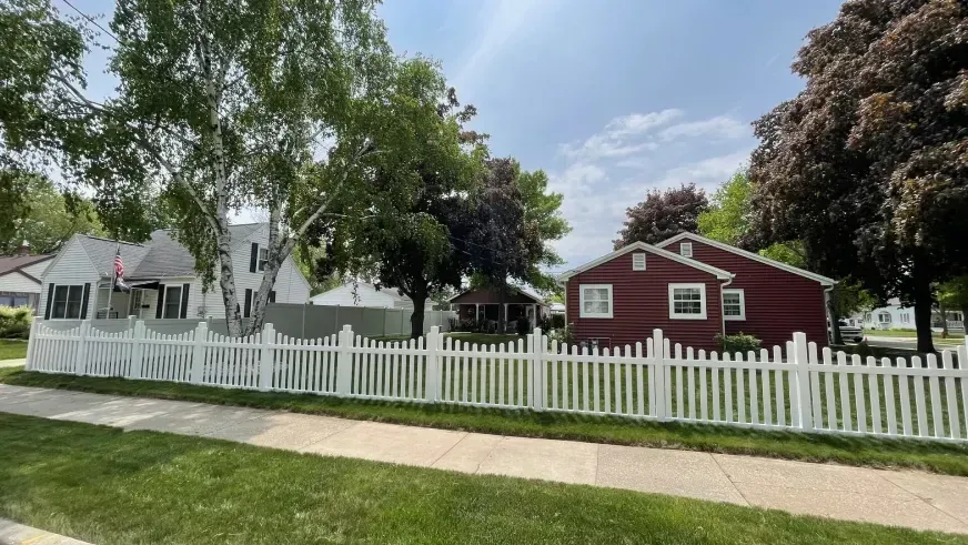 White picket fence, red house, and white tent under trees along a sidewalk on a sunny day
