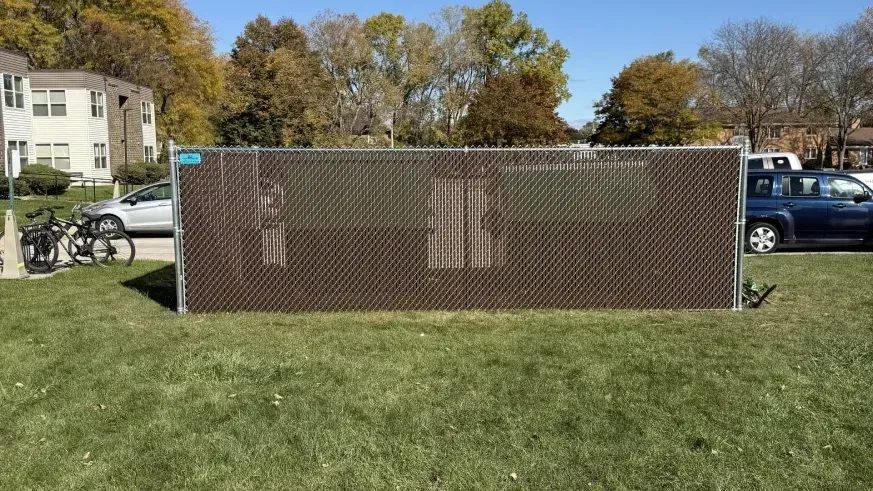Chain-link fence barrier on a grassy lawn with buildings, trees, and parked cars in the background