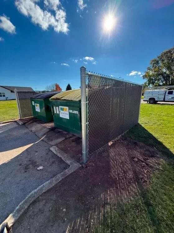 Green dumpsters beside a chain-link fence on a sunny street corner under a blue sky.