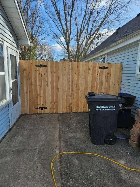 Backyard alley with a wooden fence, trash bin, brick step, and yellow hose on concrete.