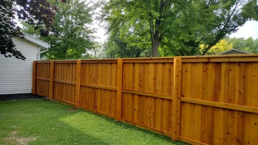 Wooden privacy fence along a green lawn beside a white house, with trees in the background.