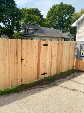 Freshly built wooden privacy fence with black hinges beside a driveway and houses.
