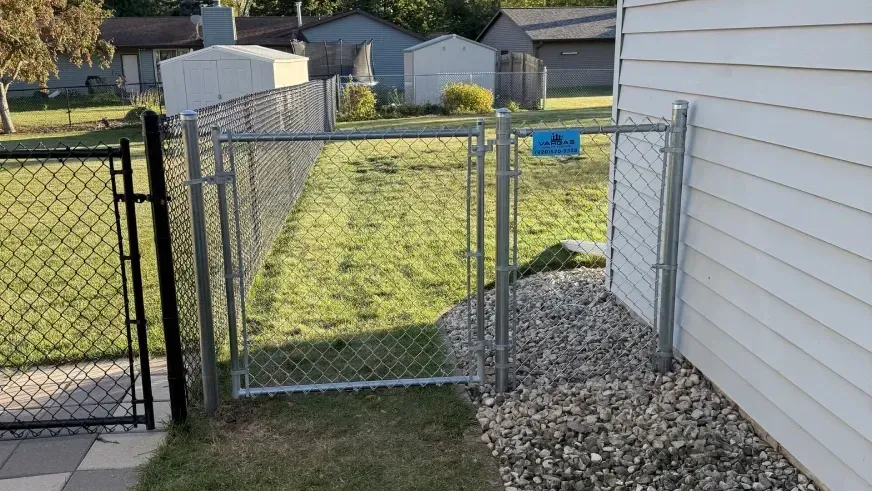 Chain-link gate beside a gravel path next to a white house, leading to a grassy backyard.