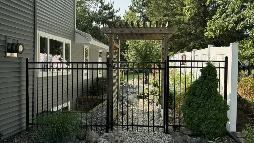 Black metal gate and pergola walkway between a gray house and white fence, with landscaped shrubs and gravel.