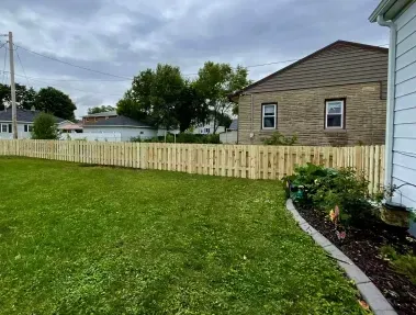 Backyard lawn with a light wooden fence, flower bed, and a house under cloudy skies