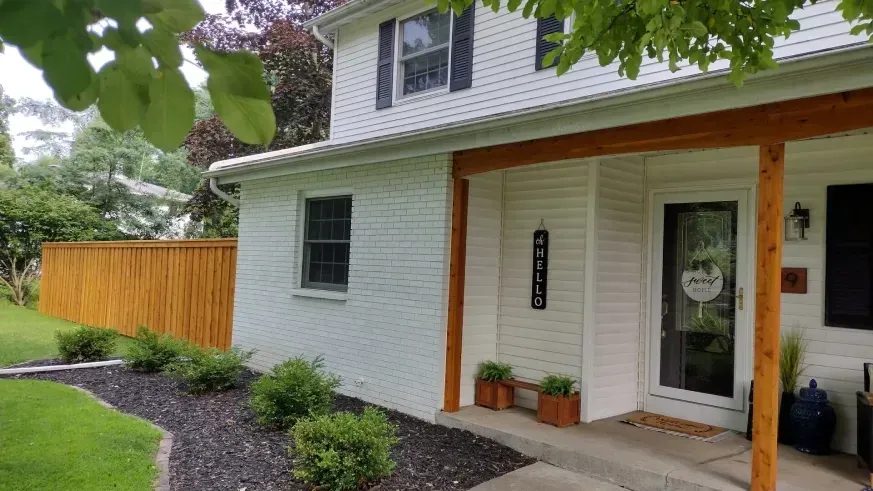 White house exterior with covered porch, wooden post, black door, and landscaped yard with fence