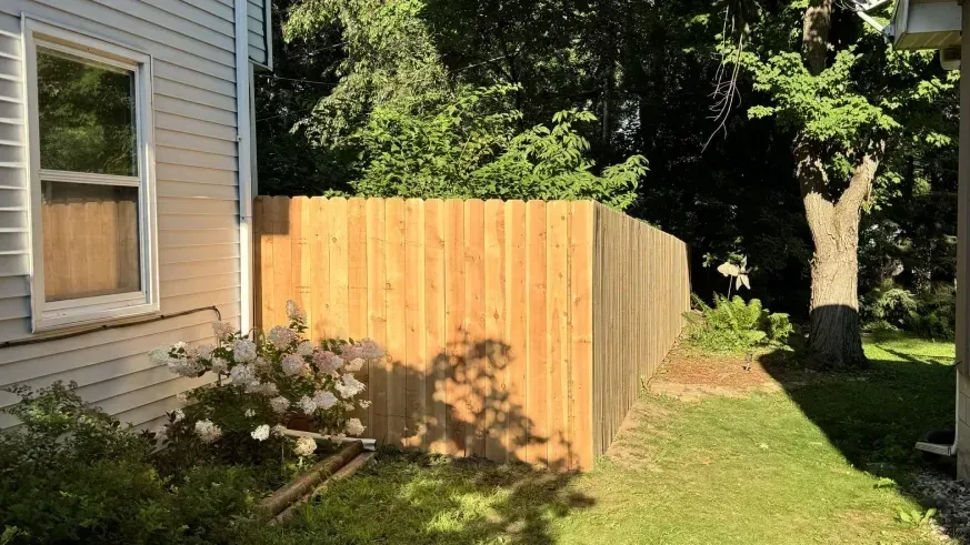 Wooden fence enclosing a narrow side yard beside a house, with trees and grass in sunlight.