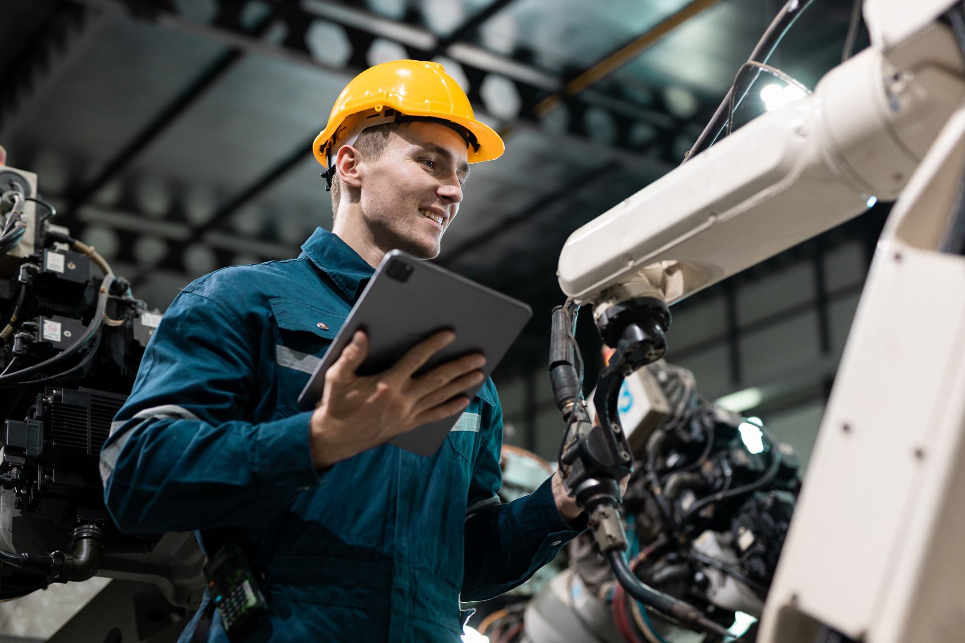 Technician in a yellow hard hat using a tablet beside industrial machinery in a factory