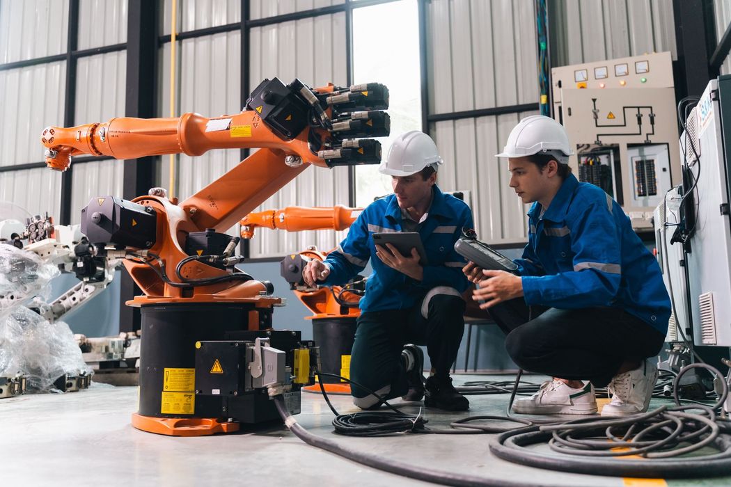 Two technicians in blue uniforms inspect a robotic arm in an industrial workshop.