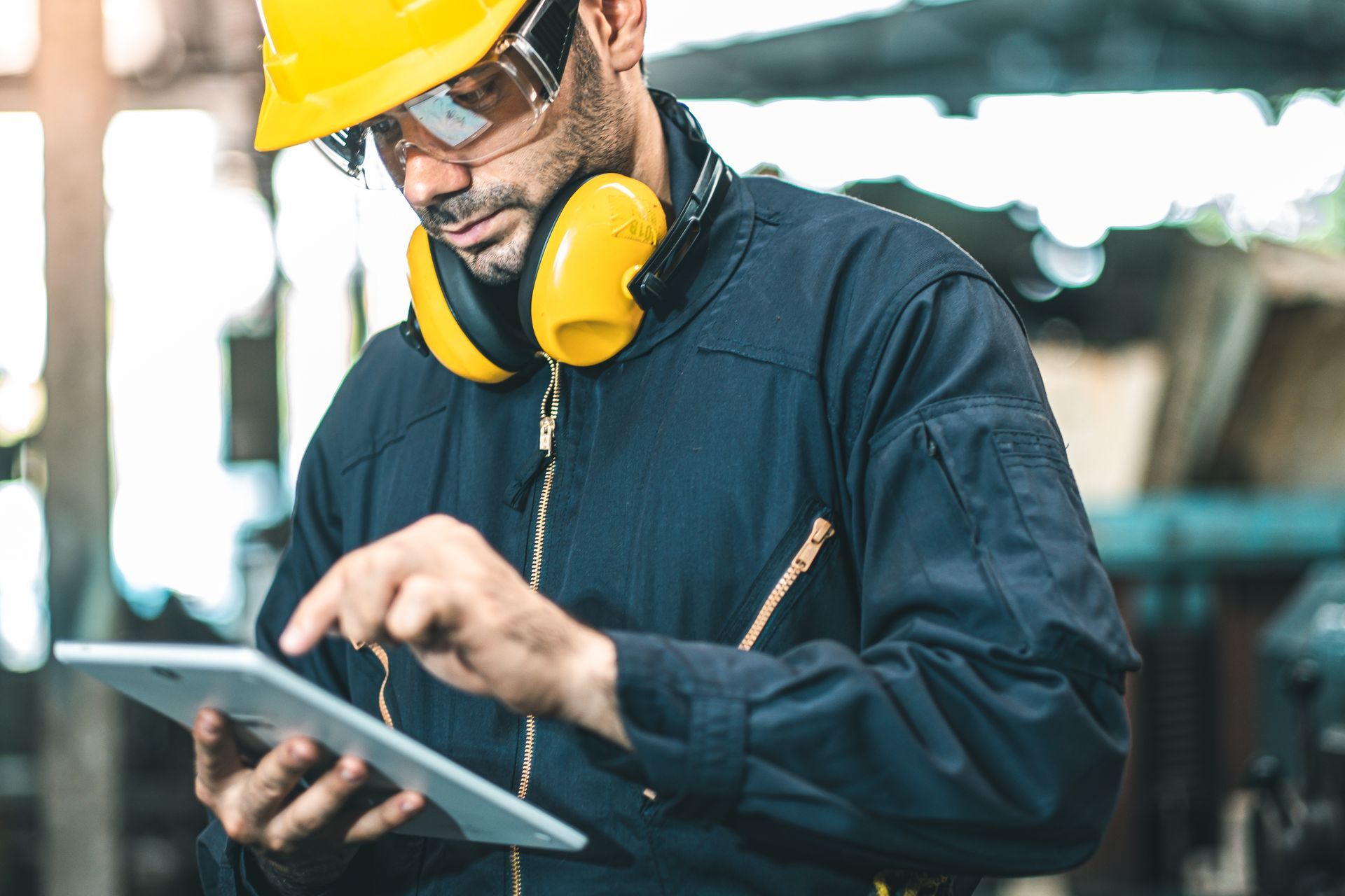 Worker in yellow hard hat and ear protection using a tablet in an industrial workshop