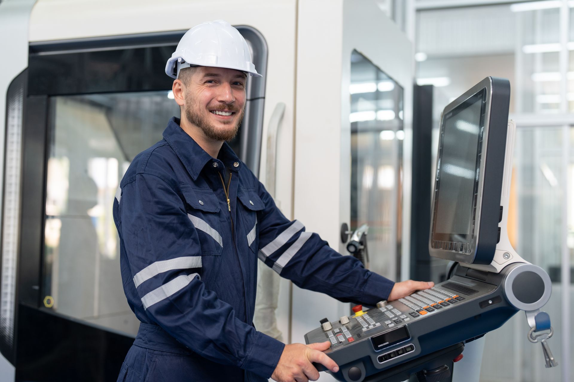 Worker in a hard hat operating a CNC machine in a factory, smiling at the control panel.