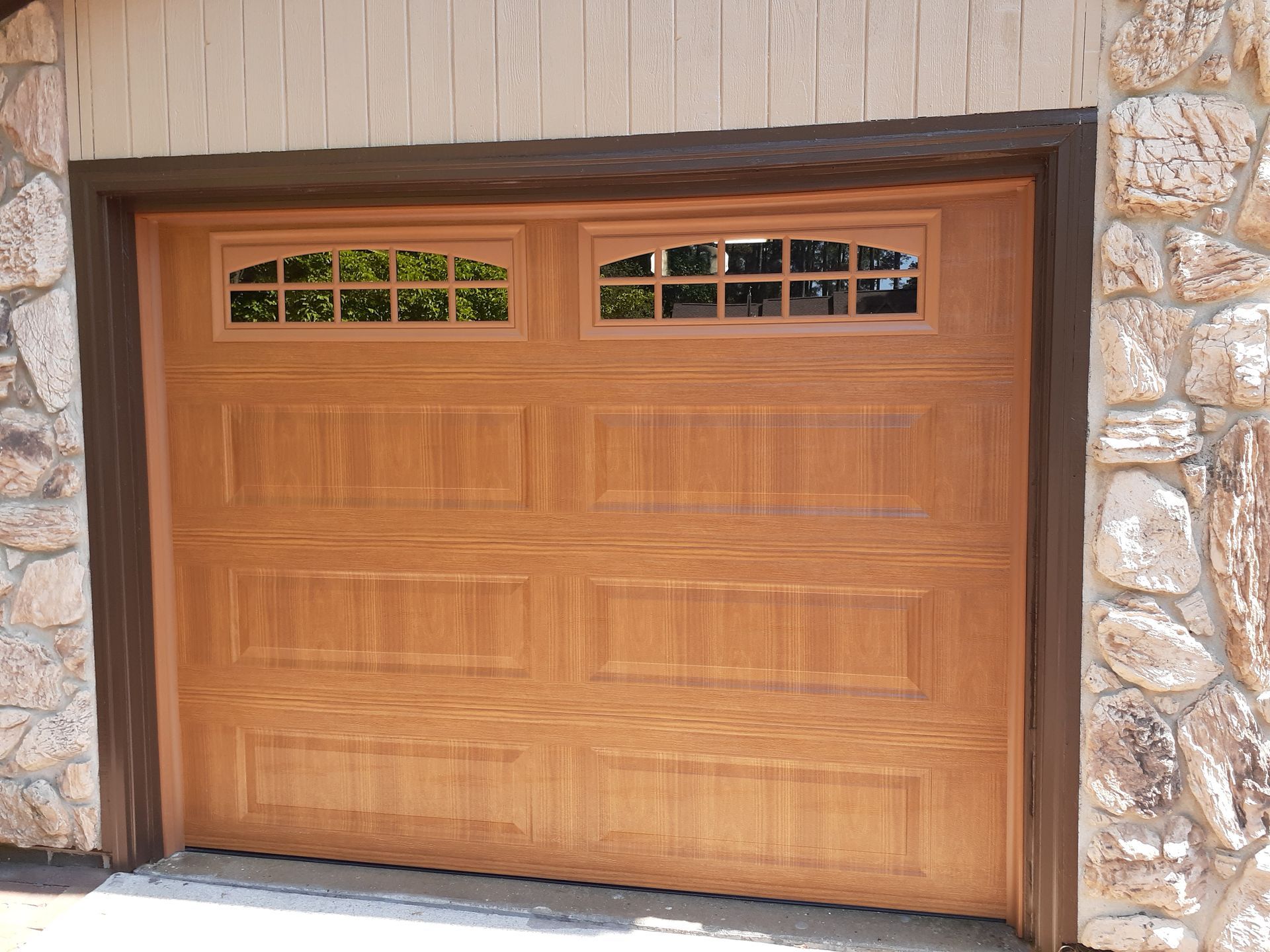 Tan garage door with two window panels above rectangular panels, set in a stone and wood frame in Lumberton, NC.