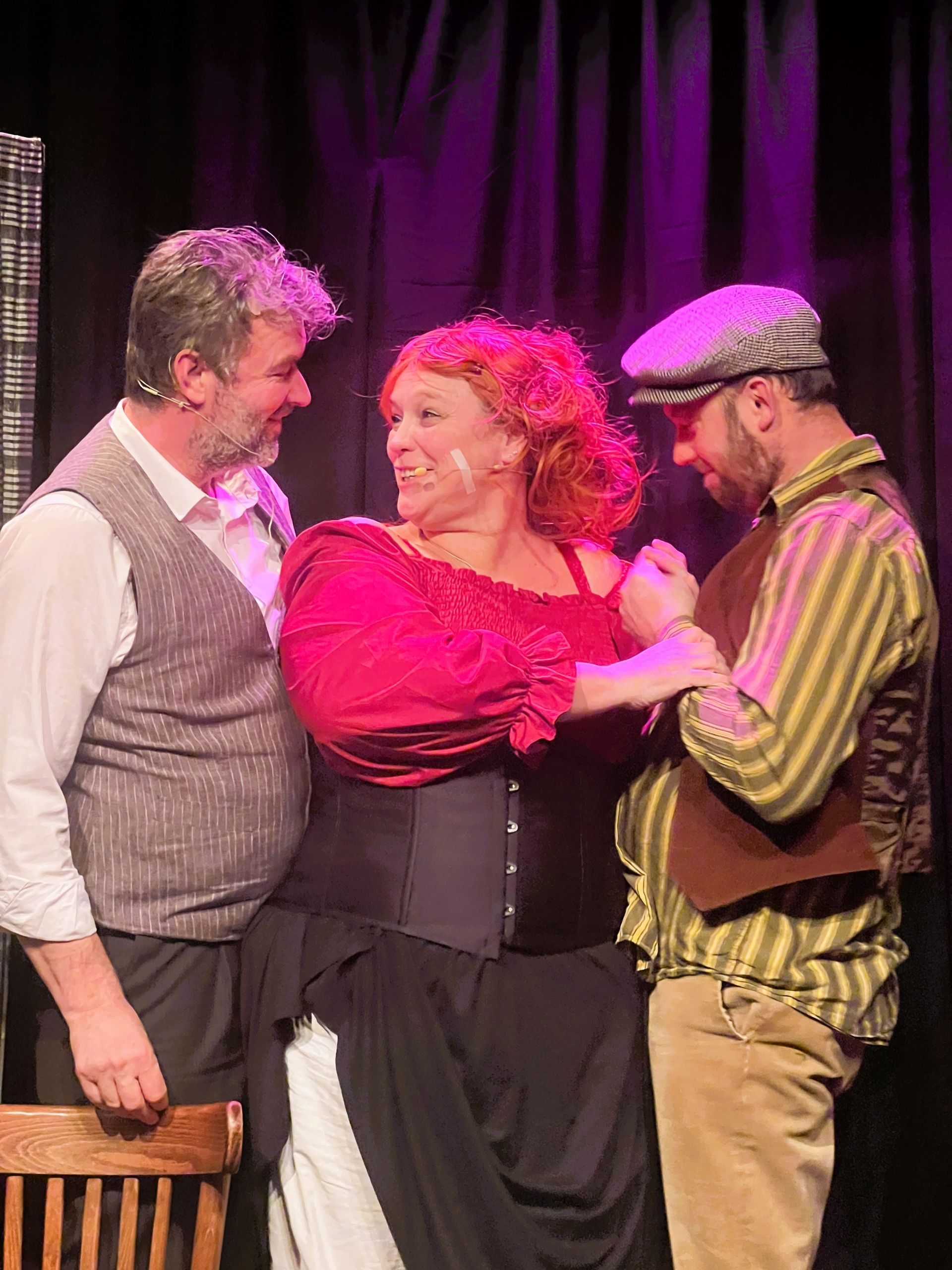 Rooie Sien. Drie acteurs in theaterkostuums op een podium. Een vrouw in rood leunt tegen twee mannen aan, die glimlachen. 