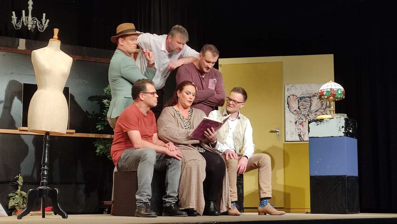 Break a leg. Een theaterpodium met zes acteurs; één leest voor, terwijl de anderen toekijken met bezorgde uitdrukkingen.