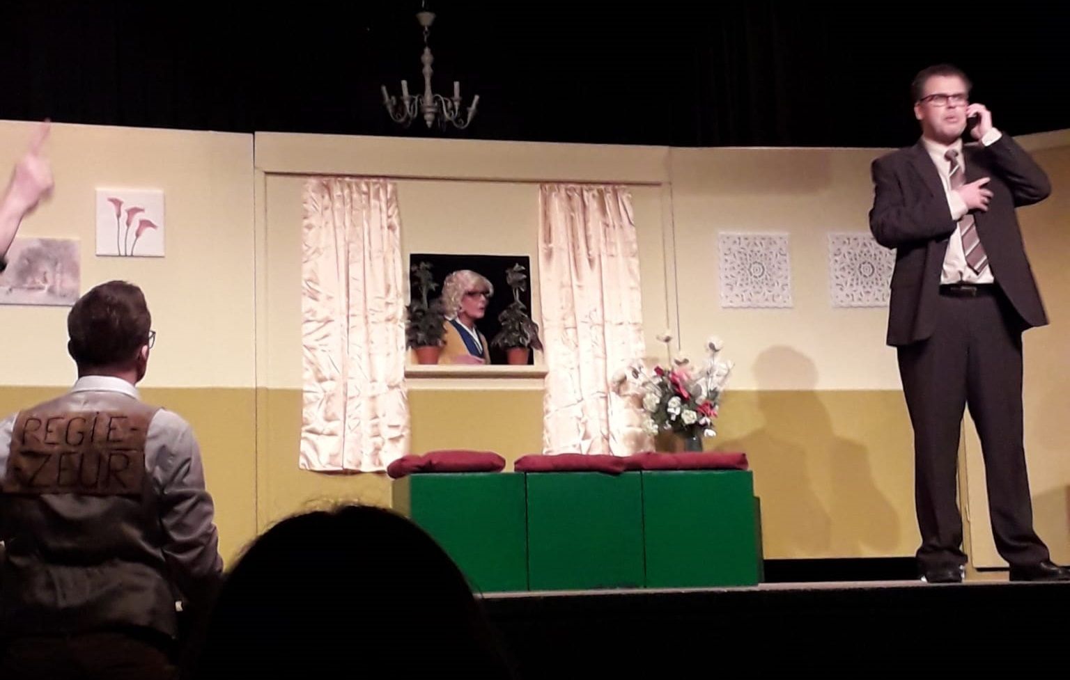Break a leg. Een toneeldecor dat een huis voorstelt. Een man belt, een ander zit, iemand kijkt uit een raam.