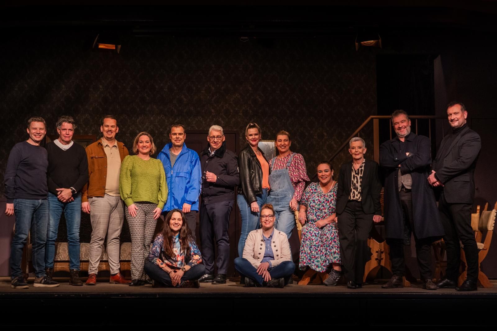 Boeven en madeliefjes. Een groep mensen staat op een podium, sommigen glimlachen. Donkere achtergrond, van bovenaf verlicht.