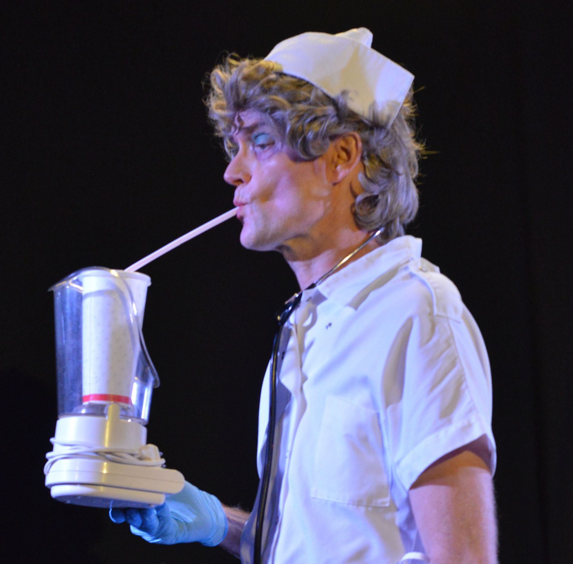 Delirium. Man in verpleegstersuniform drinkt met een rietje uit een blender, zwarte achtergrond.