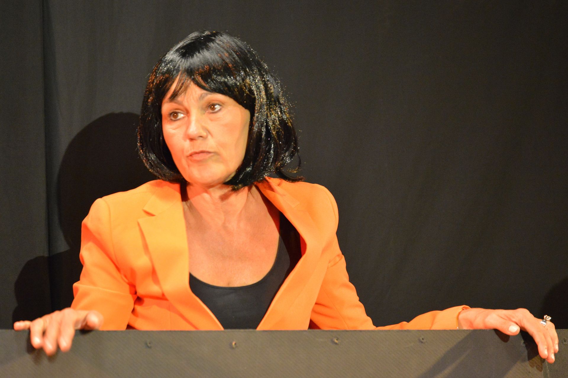 Delirium. Vrouw in een feloranje jas en zwarte pruik kijkt naar een donker oppervlak.