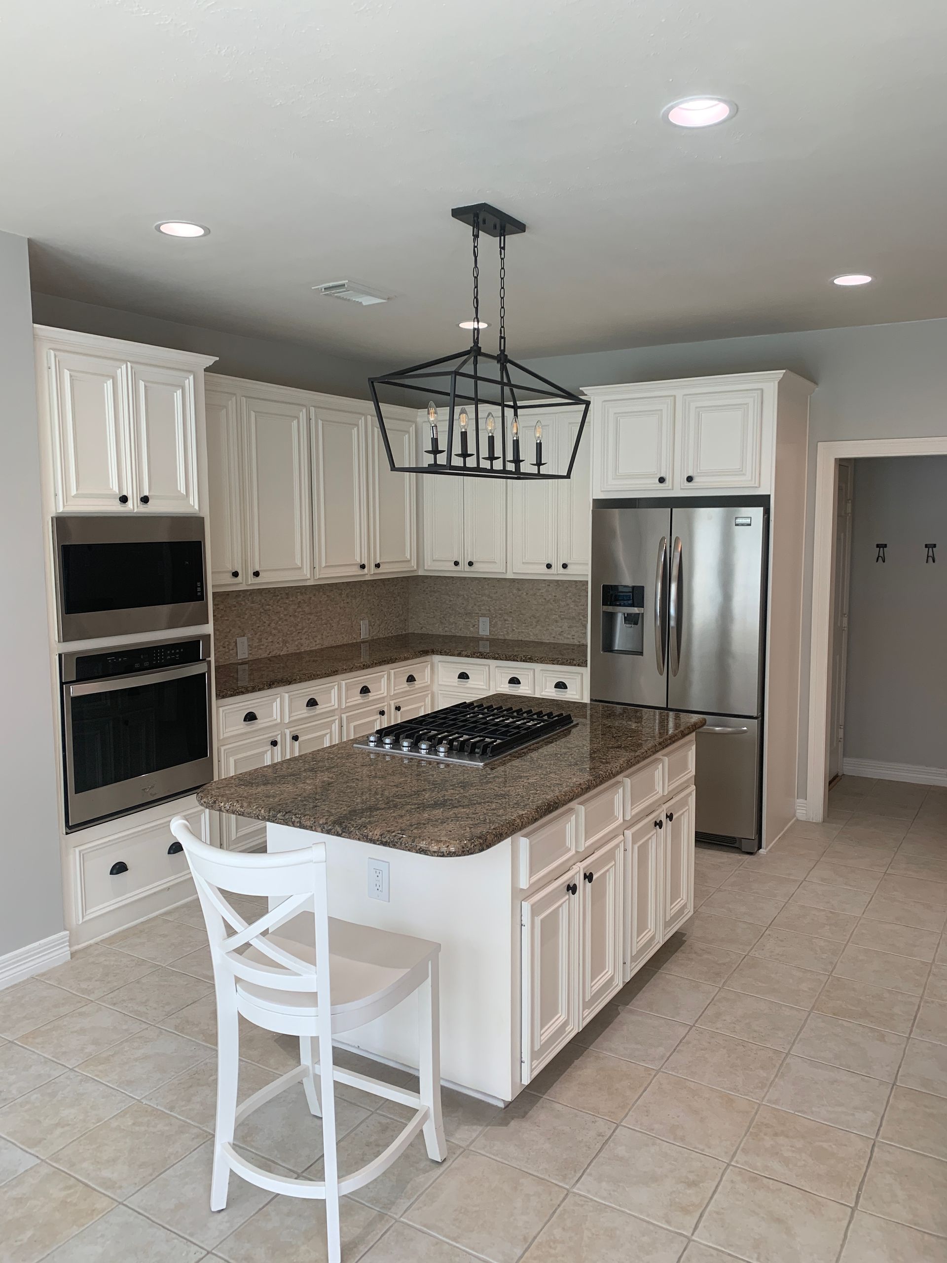 A kitchen with white cabinets, brown granite countertops, stainless steel appliances, and a central island with a chair.