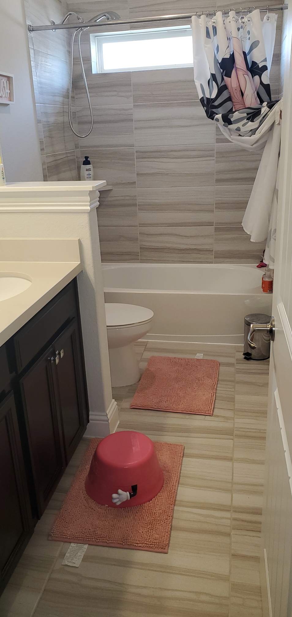 A bathroom with a dark vanity, toilet, bathtub with a patterned curtain, and two pink mats on the tile floor.