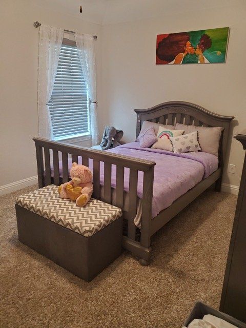 A bedroom with a gray wooden bed frame, lavender bedding, a chevron-patterned storage bench, and a colorful wall canvas.