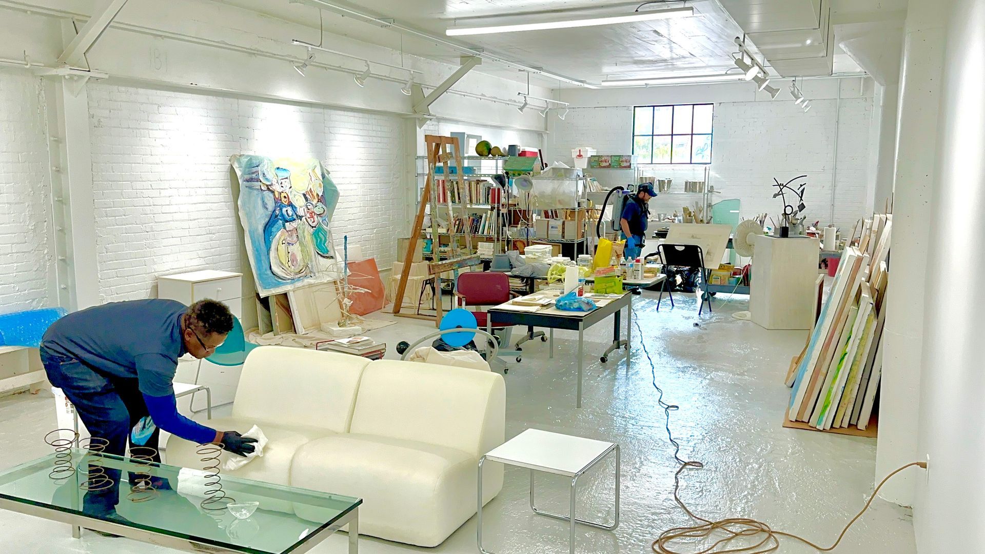 A person cleaning a white couch in a bright art studio filled with paintings, canvases, and various furniture pieces.