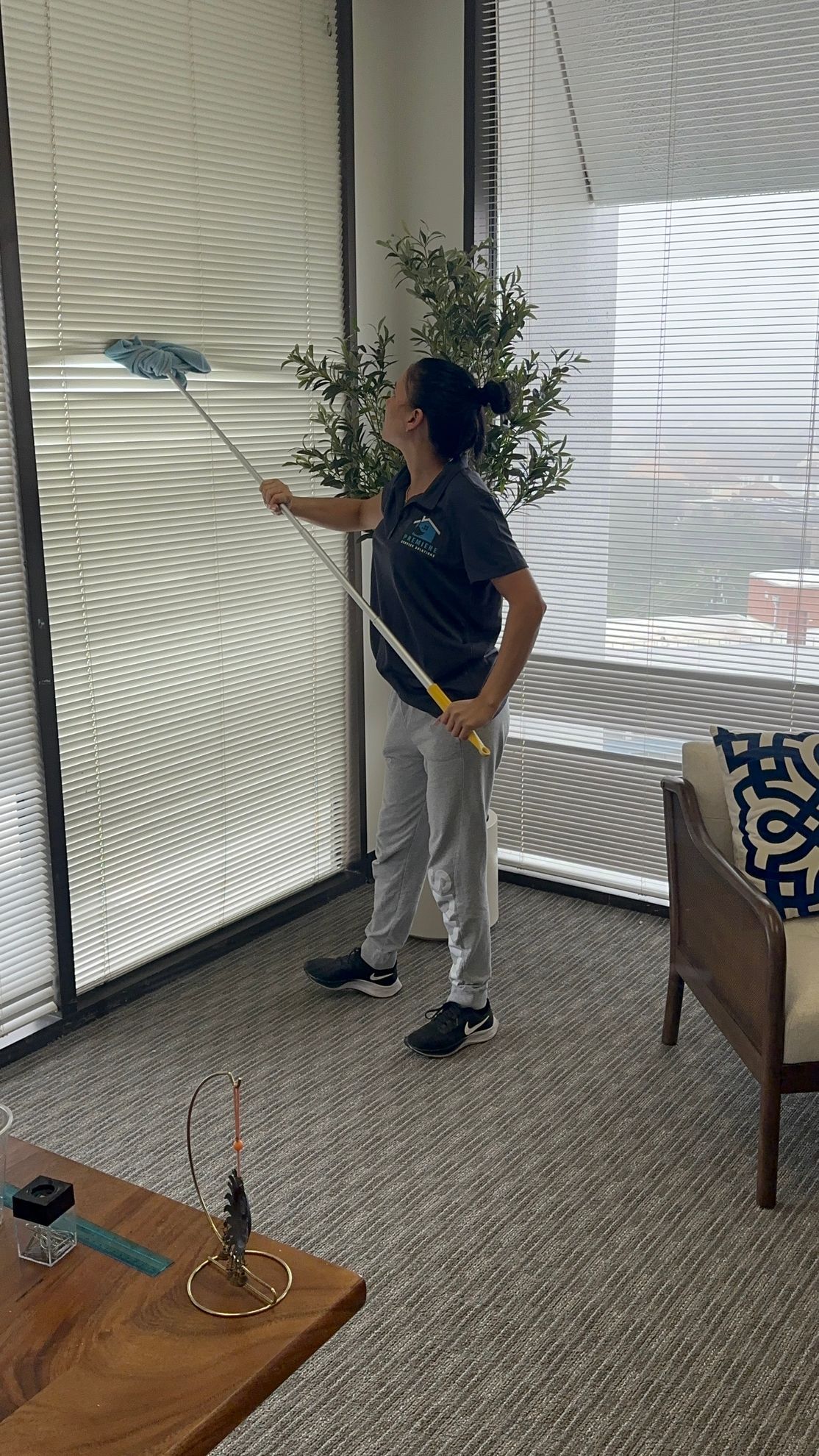 A person wearing a dark shirt and grey pants uses a long-handled duster to clean large, horizontal window blinds.