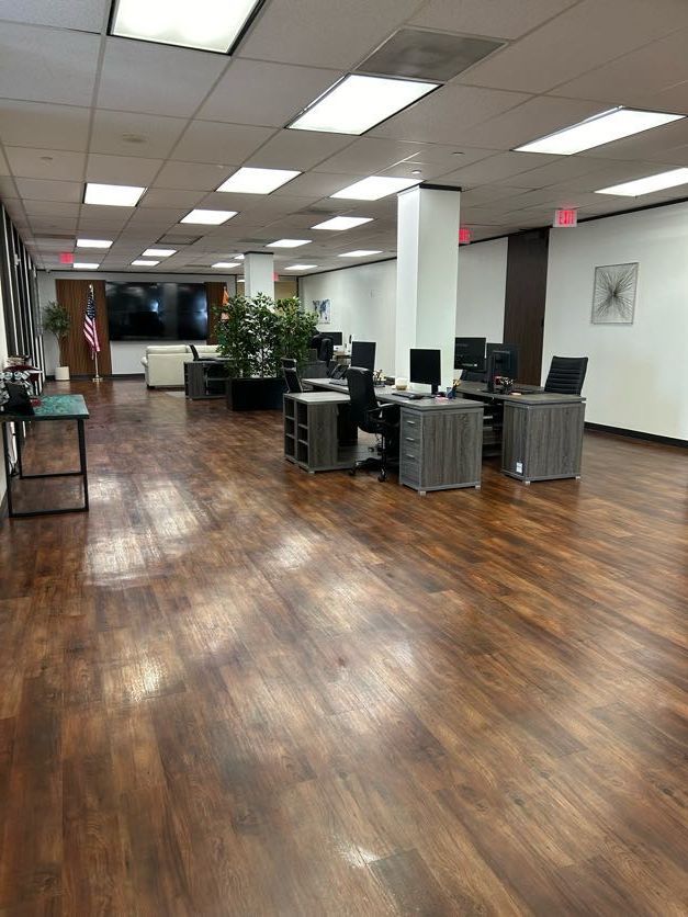 Modern office interior with wood-look flooring, multiple desks, white pillars, and large potted plants.