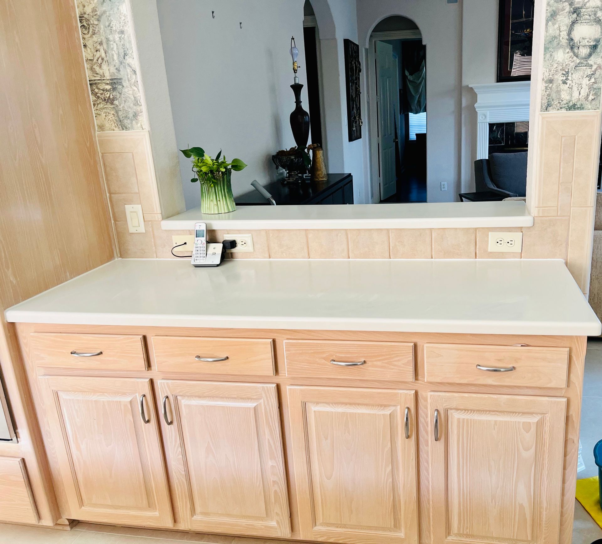 Light oak kitchen cabinets with a white countertop sit below a pass-through window looking into a home interior.